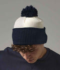 A person wearing a Beechfield Snowstar® Two Tone Beanie by Beechfield, featuring a ribbed navy blue cuff, smooth white upper section, and dark pom-pom, looks down against a plain background.