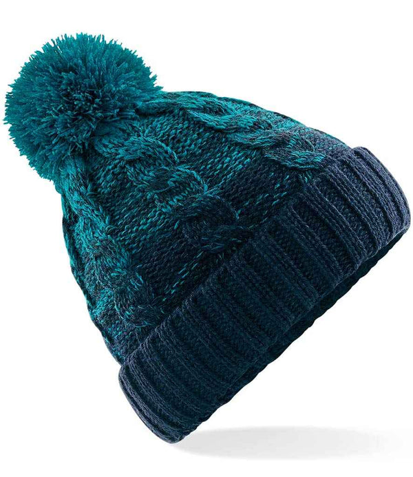 The Beechfield Ombré Pom Pom Beanie by Beechfield features blue and teal hues, a classic cable knit design, folded ribbed brim, and a large pom-pom on top.