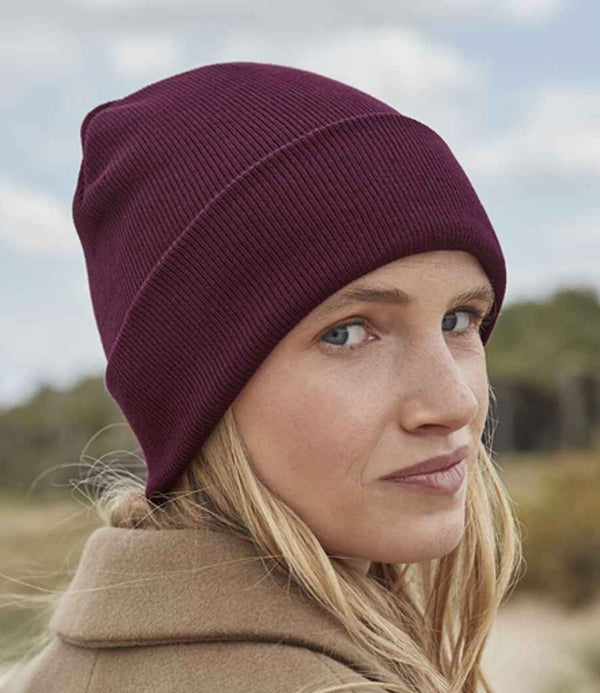 A woman with long blonde hair looks over her shoulder outdoors, wearing a maroon Beechfield Organic Cotton Original Cuffed Beanie and a tan coat. The background features greenery and a cloudy sky.