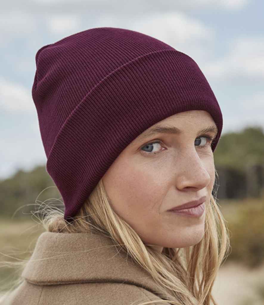 A woman with long blonde hair looks over her shoulder outdoors, wearing a maroon Beechfield Organic Cotton Original Cuffed Beanie and a tan coat. The background features greenery and a cloudy sky.
