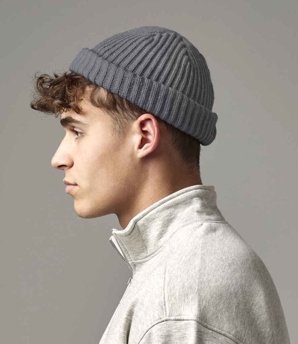 A young man with short, curly brown hair wears the Beechfield Trawler Beanie by Beechfield and a light gray zip-up sweater. He is shown in profile against a plain, neutral background.