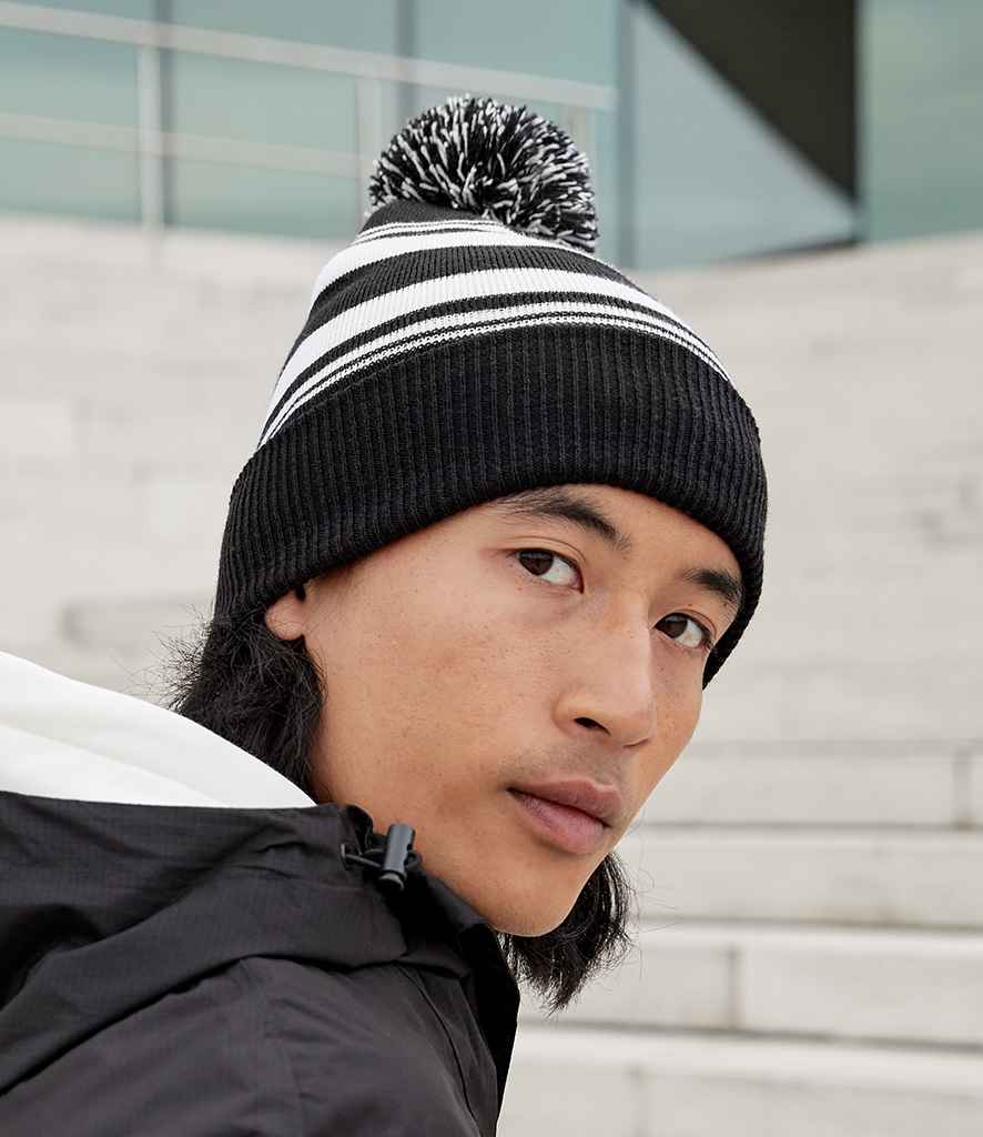 A person in a Beechfield Striped Fan Beanie and a black jacket glances over their shoulder while sitting on light-colored outdoor steps.