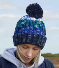 A person wearing the Beechfield Corkscrew Pom Pom Beanie by Beechfield in blue, green, and white gazes downward against a blurred outdoor background with a partly cloudy sky.