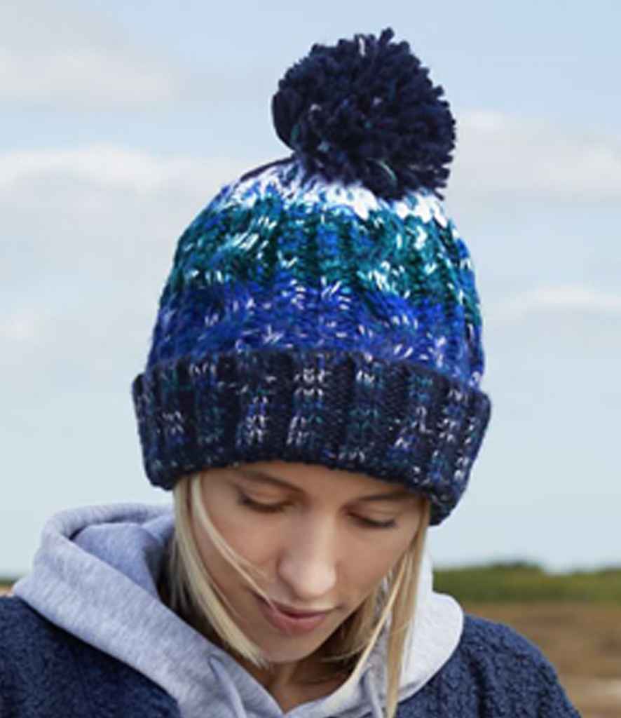 A person wearing the Beechfield Corkscrew Pom Pom Beanie by Beechfield in blue, green, and white gazes downward against a blurred outdoor background with a partly cloudy sky.