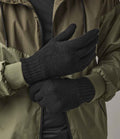 A person wearing a two-tone green jacket with ribbed cuffs and black Beechfield Classic Thinsulate™ Gloves, one gloved hand adjusting the other, stands against a plain neutral background.