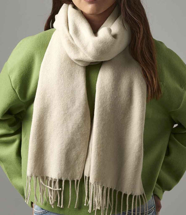 A person wears a light green sweater and the Beechfield Classic Woven Scarf, a cream-colored accessory with an oversized design and fringed tassel trim by Beechfield, wrapped around the neck. The persons face is partially out of frame.