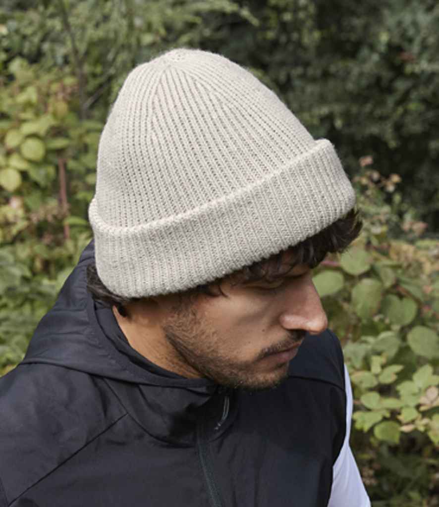 A person in a light-colored Beechfield Recycled Wind Resistant Breathable Elements Beanie with a cuffed design and dark jacket stands outdoors before green foliage, looking down and slightly to the side.