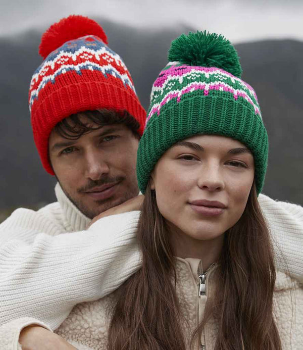A man and a woman wear Beechfield Retro Ski Beanies outdoors—his in red with a Fair Isle pattern, hers in green with pink and white designs. Both pair the beanies with cream jackets against a mountain backdrop. Brand: Beechfield.