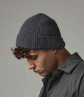 A young man with short curly hair looks downward wearing a Beechfield Organic Cotton Waffle Beanie in dark gray and a dark gray button-up shirt, set against a plain gray background.