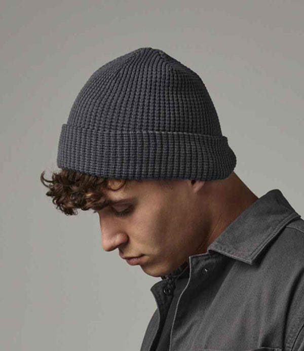 A young man with short curly hair looks downward wearing a Beechfield Organic Cotton Waffle Beanie in dark gray and a dark gray button-up shirt, set against a plain gray background.