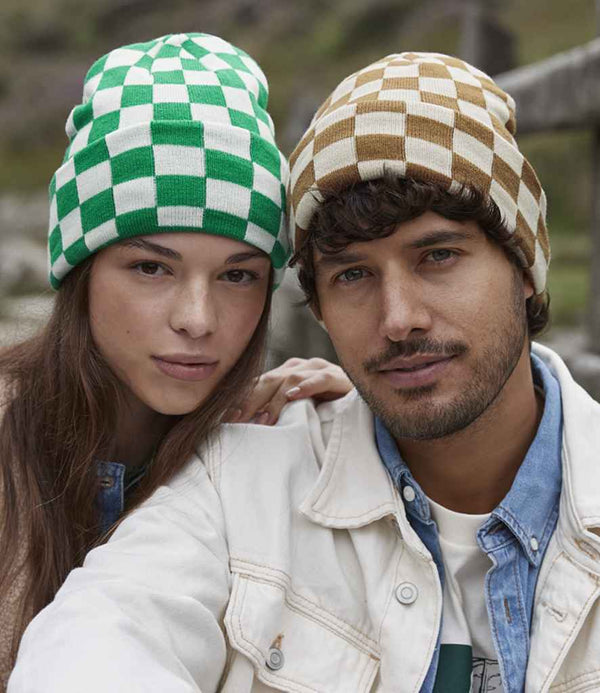 Two people pose closely outdoors. The left wears a Beechfield Checkerboard Beanie in green and white, while the right sports a brown and beige wide cuffed beanie paired with a light-colored jacket.