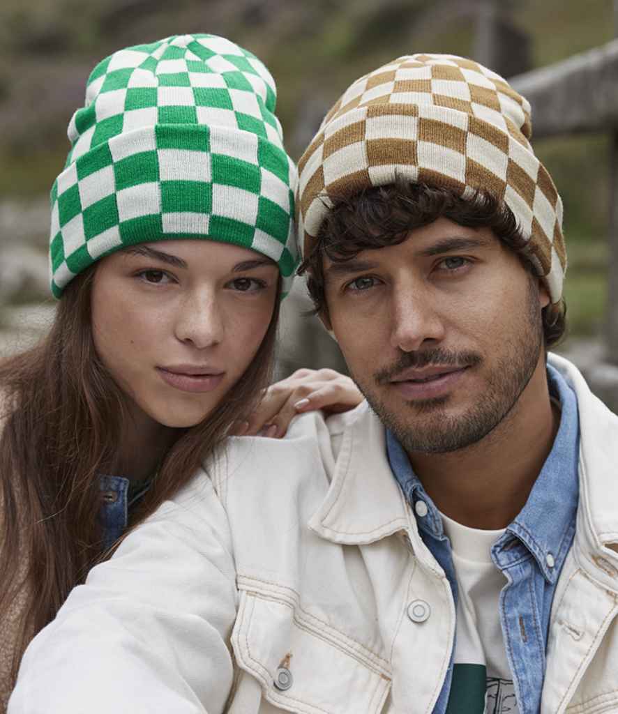Two people pose closely outdoors. The left wears a Beechfield Checkerboard Beanie in green and white, while the right sports a brown and beige wide cuffed beanie paired with a light-colored jacket.