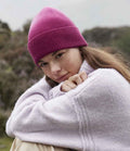 A person in a Beechfield Dip Dye Beanie by Beechfield and a light purple sweater sits outdoors with arms around their knees, looking at the camera. The background features blurred greenery.