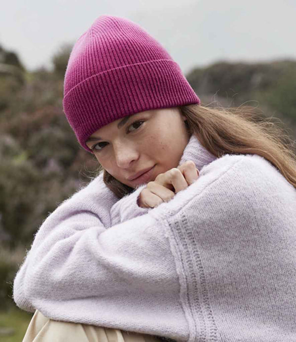 A person in a Beechfield Dip Dye Beanie by Beechfield and a light purple sweater sits outdoors with arms around their knees, looking at the camera. The background features blurred greenery.