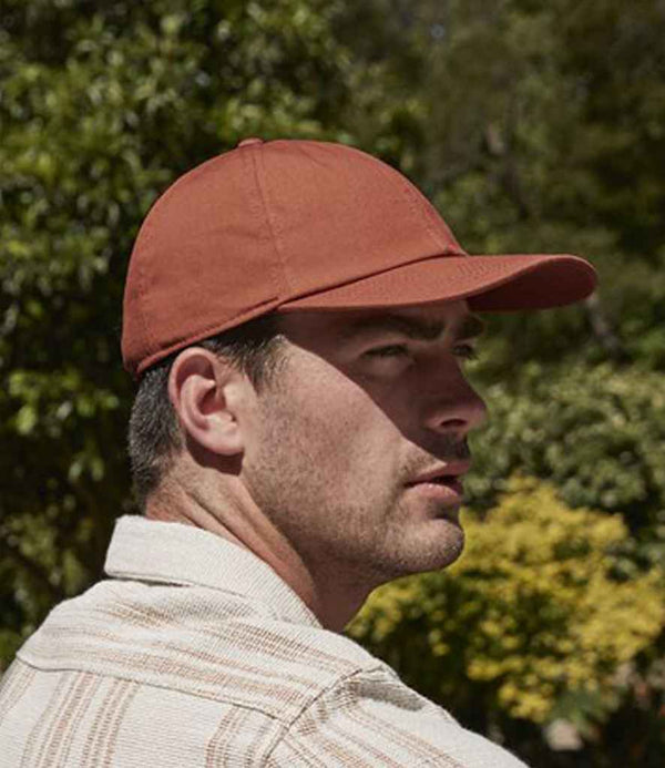 A man wearing a Beechfield Organic Cotton 6 Panel Cap in brown and a light-striped shirt looks to his right outdoors, with green trees and shrubs in the background.