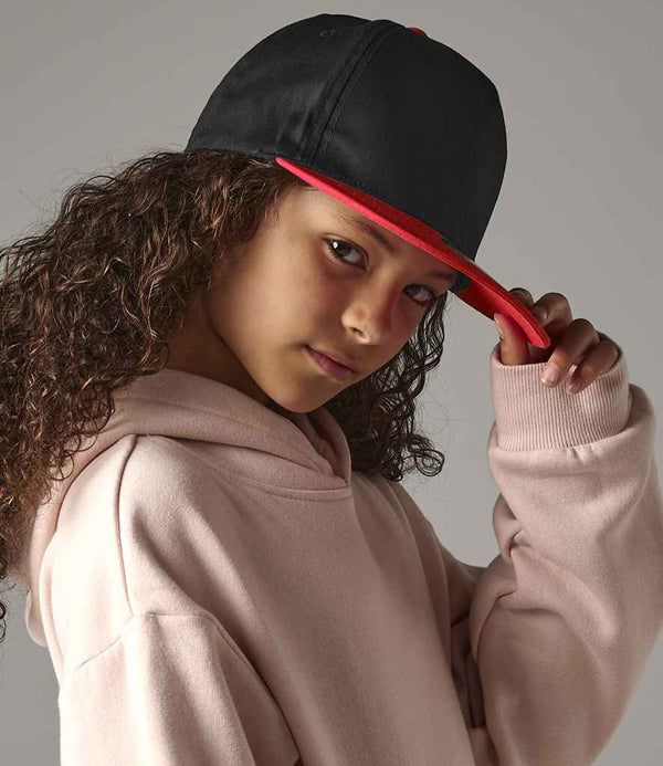 A person with long curly hair wears a light pink hoodie and tilts a black and red Beechfield Youth Snapback Cap by Beechfield forward, looking at the camera against a plain background.