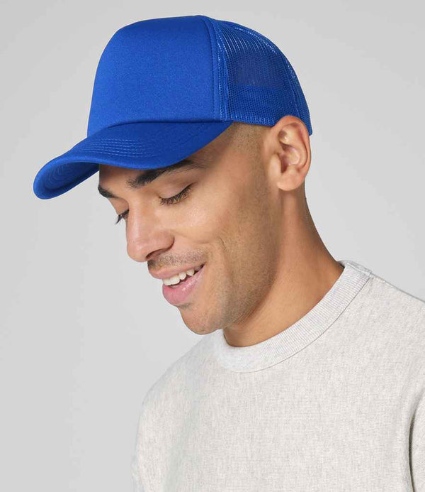 A person in a Beechfield Classic Foam Trucker Cap by Beechfield, featuring bright blue retro mesh-back and snapback closure, and a light gray crewneck sweatshirt, smiles slightly while looking down against a plain light background.