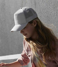 A person with long hair wears a light Beechfield Kids Snapback Trucker Cap by Beechfield and a striped shirt, standing outdoors by a textured gray wall and looking down as shadows fall across the surface.