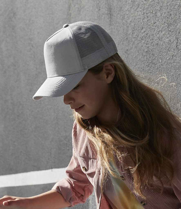 A person with long hair wears a light Beechfield Kids Snapback Trucker Cap by Beechfield and a striped shirt, standing outdoors by a textured gray wall and looking down as shadows fall across the surface.