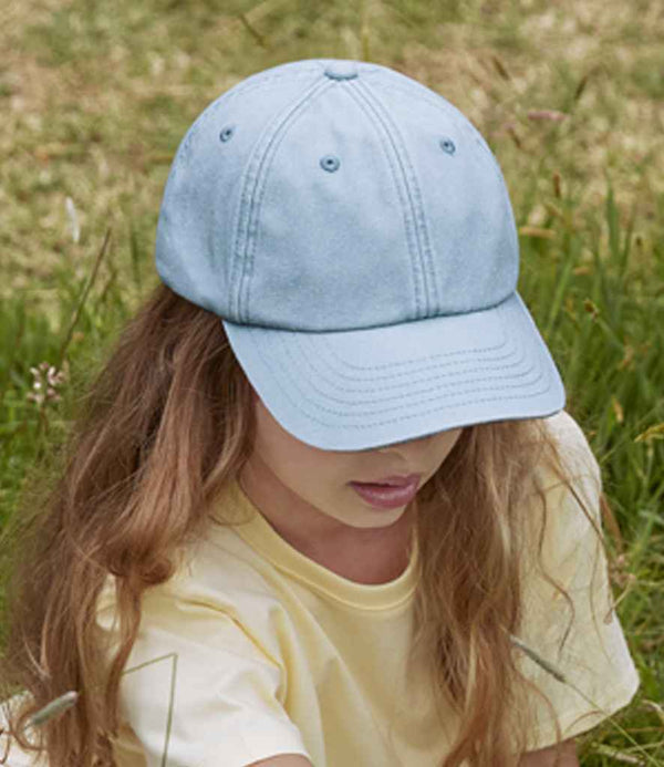 A person with long light brown hair wearing a pale yellow shirt and a Beechfield Junior Low Profile Vintage Cap is sitting outdoors on grass, the cap’s brim covering most of their face.
