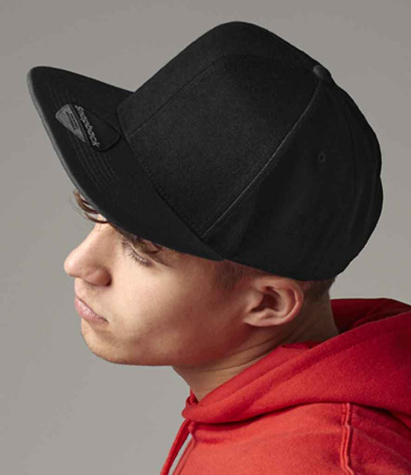 A young person in profile wears a Beechfield Original Flat Peak 6 Panel Snapback Cap and a red hoodie against a plain background, looking slightly downward with a neutral expression.