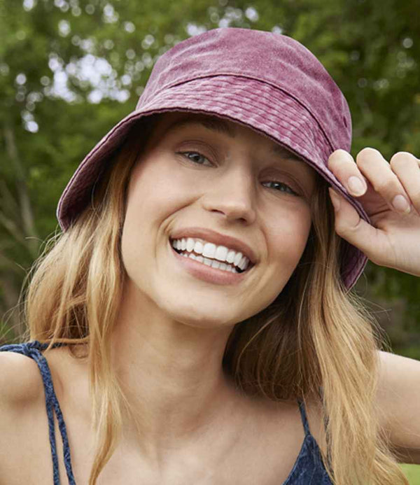 A person with long blonde hair smiles at the camera, wearing a sleeveless top and a maroon Beechfield Vintage Bucket Hat by Beechfield for sun protection, holding the brim. Green trees appear blurred in the background.