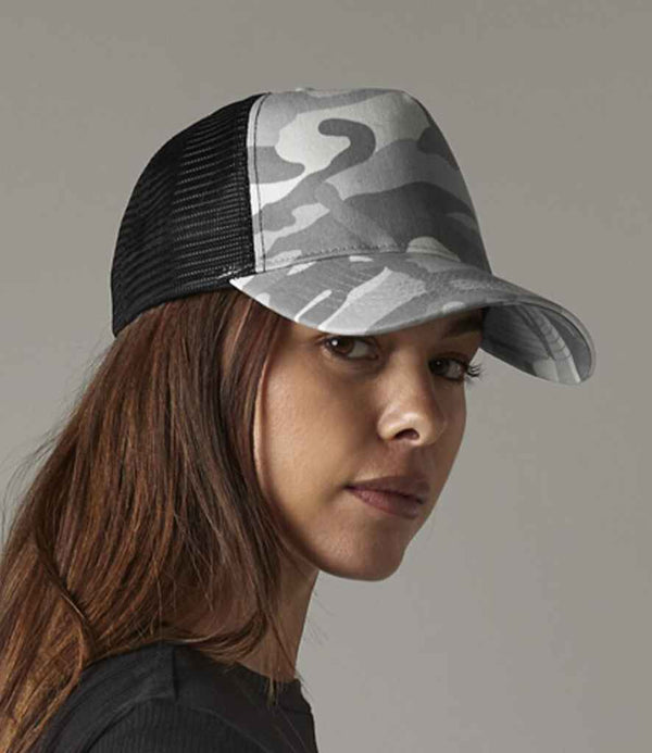A woman with long brown hair wears a Beechfield Camo Snapback Trucker Cap with a polyester mesh back and a black shirt, looking over her shoulder against a plain background.