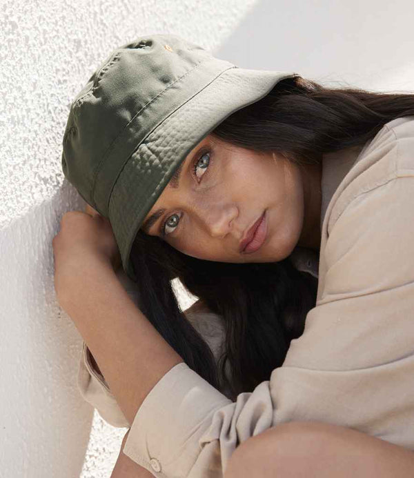 A woman with long dark hair, wearing the Beechfield Recycled Polyester Bucket Hat in khaki and a beige shirt, sits against a white textured wall and gazes directly at the camera.