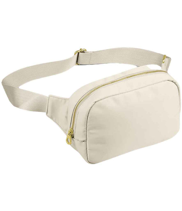 The BagBase Sundae Cross Body Bag by BagBase is a beige fanny pack with premium fabric, a zippered main compartment, adjustable webbing strap, and gold-tone zipper hardware, shown on a white background.