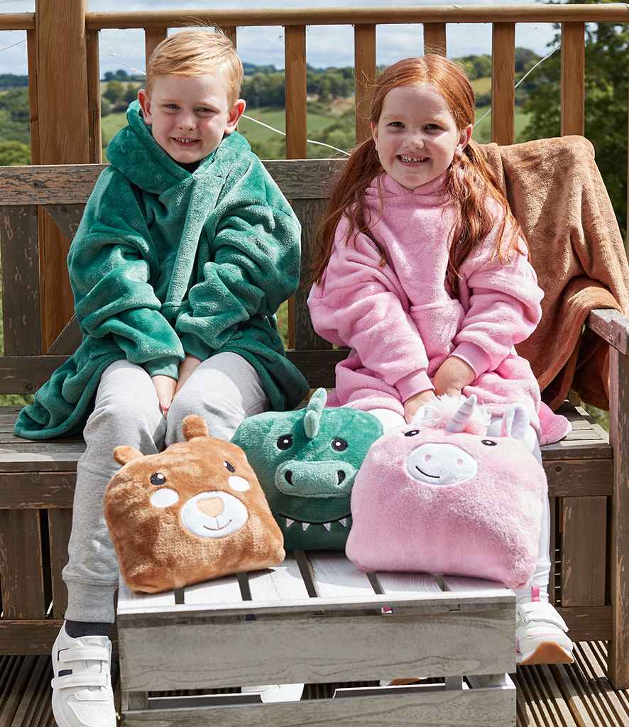 Two kids sit outdoors on a wooden bench, both wearing oversized Brand Lab Kids Character Cushion Hooded Blankets—one green, one pink. In front of them are three Brand Lab character soft toys: a bear, dinosaur, and unicorn.
