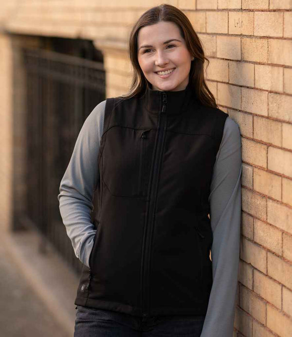A woman with long brown hair smiles while leaning against a beige brick wall, wearing the Stormtech Ladies Cascades Soft Shell Bodywarmer over a gray long-sleeve shirt, hands in her pockets.