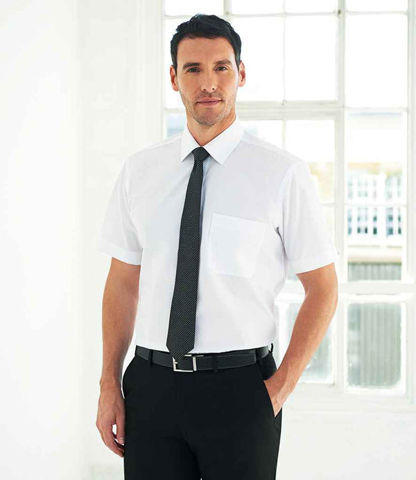 A man stands indoors, hands in pockets, wearing the Brook Taverner Rosello Short Sleeve Poplin Shirt in white with a black tie and trousers. He faces forward; large windows fill the background with natural light. Brand: Brook Taverner.