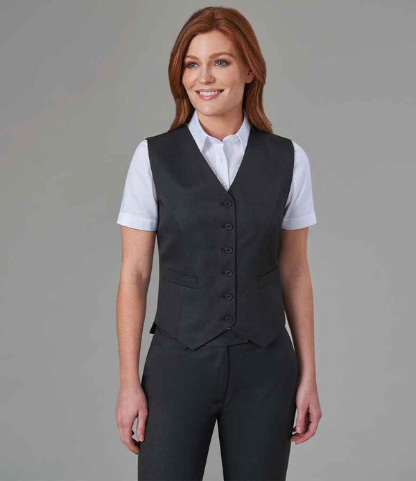 A woman with long red hair smiles in front of a gray background, wearing the Brook Taverner Ladies Concept Omega Waistcoat— a dark gray single-breasted vest from Brook Taverner — over a white short-sleeve button-up shirt with matching pants.