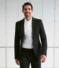 A man in a Brook Taverner Jersey Stretch Rory Jacket, white dress shirt, and black pants stands indoors smiling at the camera. White panels with rectangular outlines appear in the background.