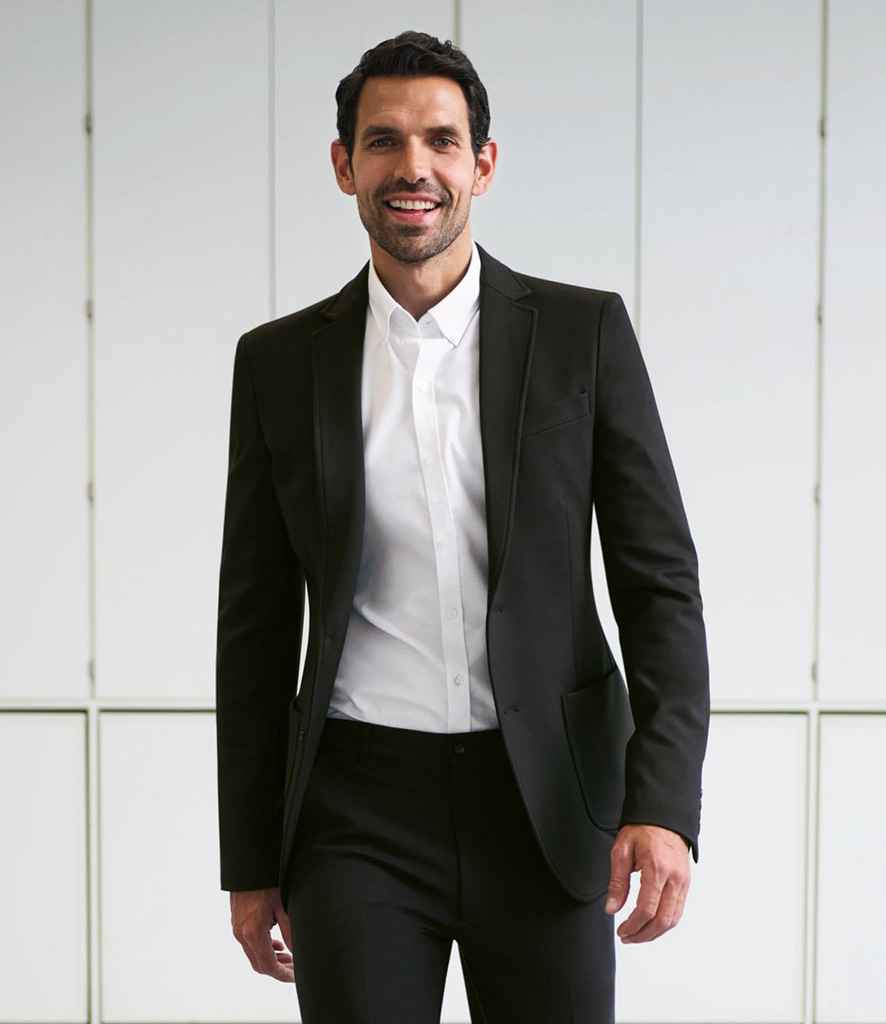A man in a Brook Taverner Jersey Stretch Rory Jacket, white dress shirt, and black pants stands indoors smiling at the camera. White panels with rectangular outlines appear in the background.