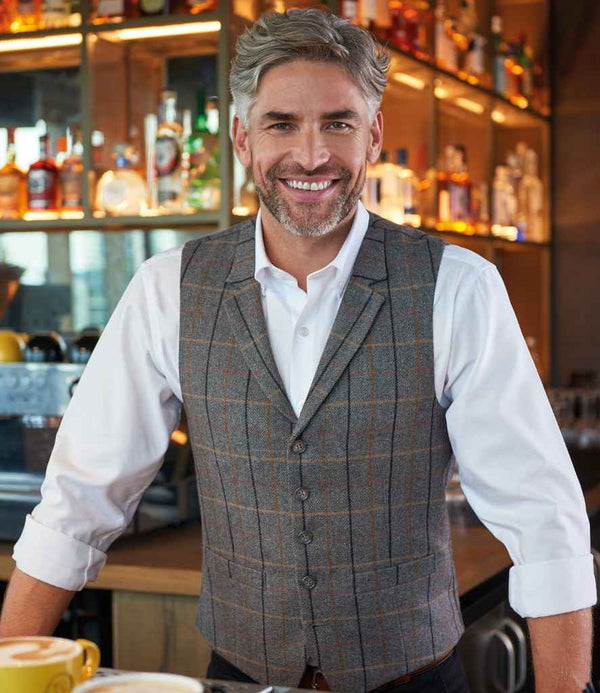 A gray-haired, bearded man smiles behind a bar, wearing a white shirt and the Brook Taverner Memphis Tweed Waistcoat. Shelves with bottles and glasses are visible in the background.