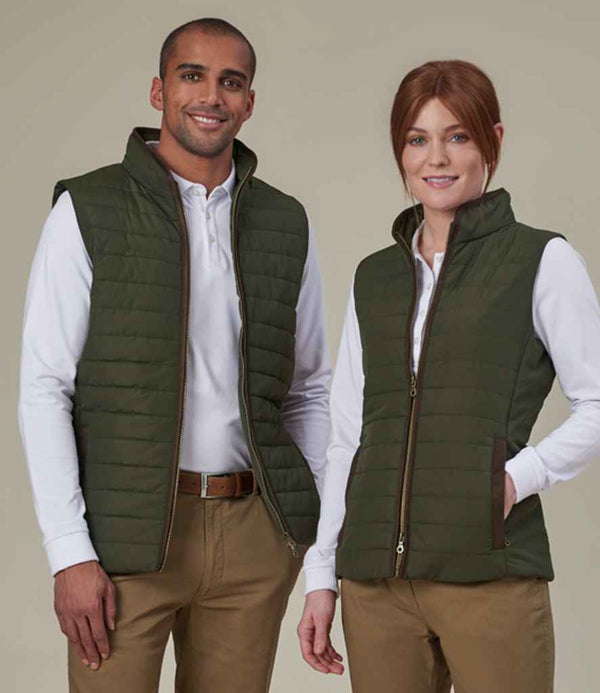 Two people stand side by side against a beige background, both wearing Brook Taverner Ladies Madison Quilted Gilets in olive green over white long-sleeve shirts and khaki pants. The vests feature showerproof recycled polyester and Repreve recycled padding.