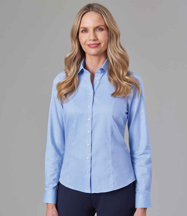A woman with long blonde hair wears the Brook Taverner Ladies Aspen Long Sleeve Oxford Shirt, a light blue tailored fit with a semi-cut away collar, paired with dark pants. She stands against a plain gray background, facing the camera with a slight smile.