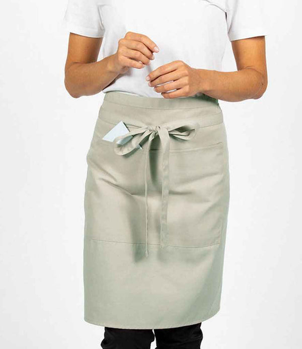 A person wears a white short-sleeve shirt and a Dennys Polyester Waist Apron with Pocket in light gray, featuring a crease-resistant finish, front pocket holding a piece of paper, and tied with a bow. Their hands are raised.