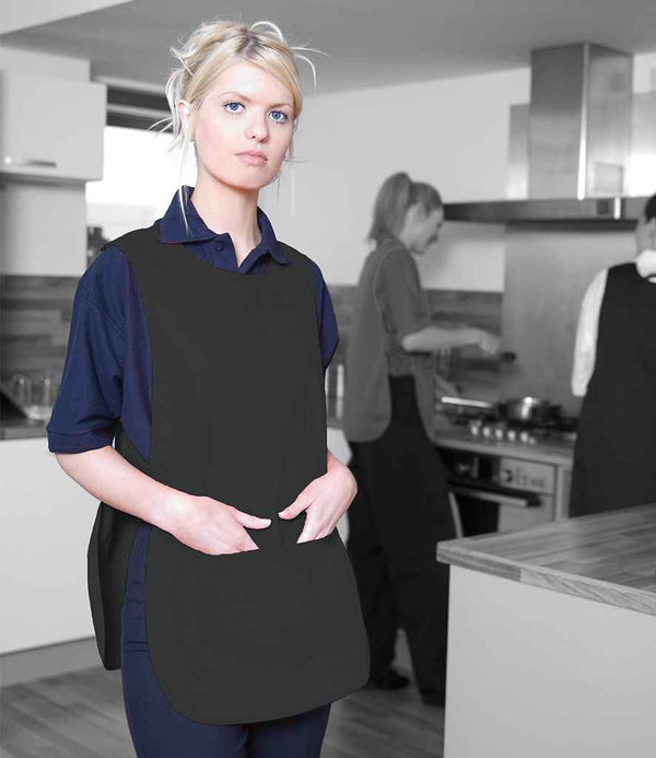 A woman wearing a Warrior Pocket Tabard by Warrior with adjustable side tabs stands in a kitchen, facing the camera, while two others in matching tabards prepare food at the stove and counter in the background.