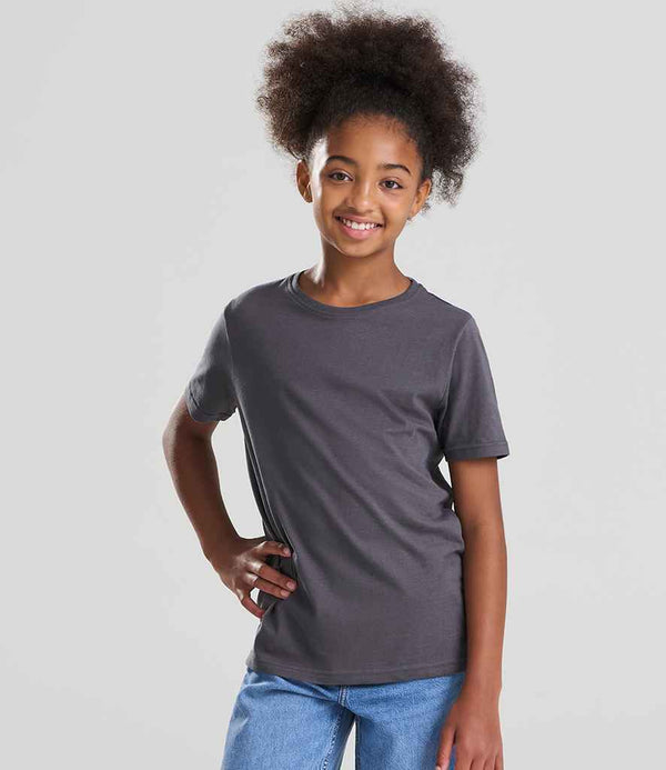 A young girl smiles confidently with one hand on her hip, wearing blue jeans and the Ecologie Kids Cascades Organic T-Shirt by Ecologie—a dark gray, fashion fit top with a ribbed collar—against a light gray background. Her curly hair is pulled back.