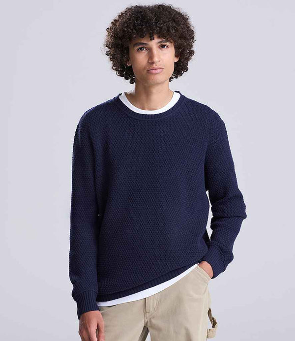 A person with curly hair models the Ecologie Unisex Taroko Sustainable Crew Neck Sweater in dark blue, made from recycled cotton, over a white shirt and beige pants, standing against a plain light gray background.
