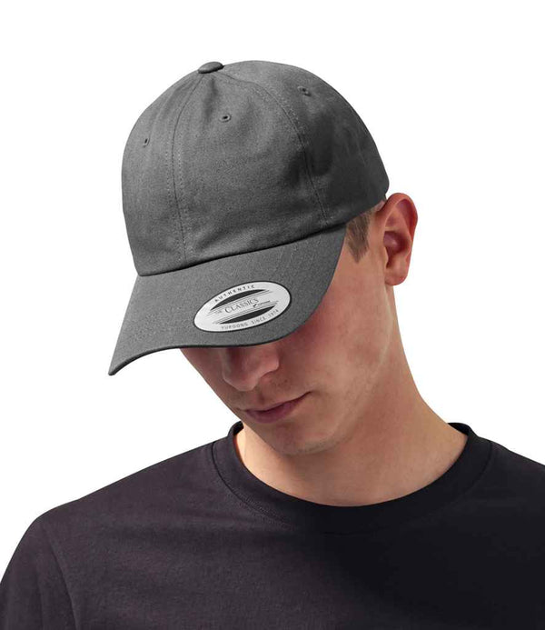 A person in a Flexfit Low Profile Cotton Twill Cap in dark gray, featuring a pre-curved peak and CLASSIC sticker, pairs it with a black shirt against a plain white background while looking downward.