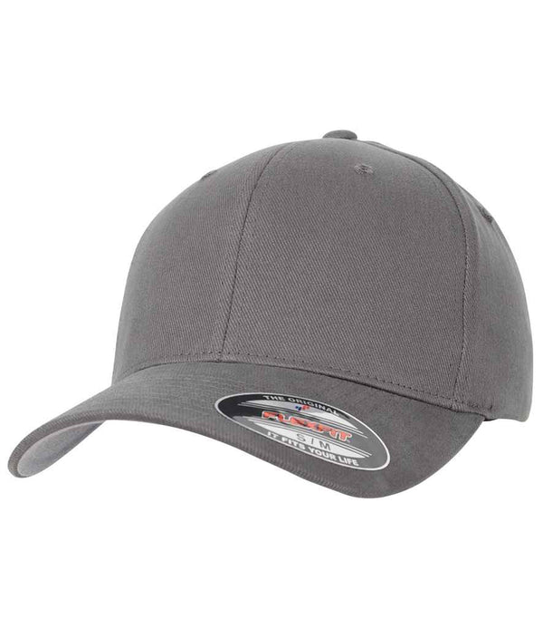 The Flexfit Brushed Twill Cap by Flexfit is a plain gray 6-panel cap with a curved peak and structured front panels. A sticker on the bill shows product details. The cap is shown against a white background.