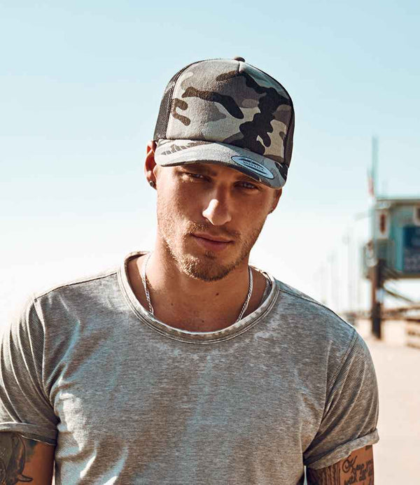A man wearing a Flexfit Classic Camo Trucker Cap by Flexfit and a gray t-shirt stands outside on a sunny day. He has arm tattoos, a neutral expression, and there’s a blurred structure in the background.