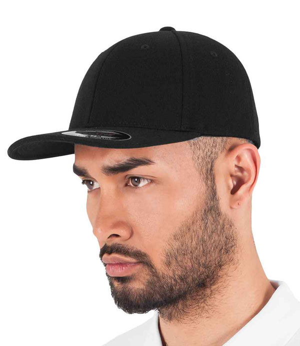 A man with a trimmed beard wears a black Flexfit Double Jersey Cap by Flexfit, featuring a mid profile and flat brim with a small sticker under the bill. He is dressed in a white shirt and gazing slightly to the side.