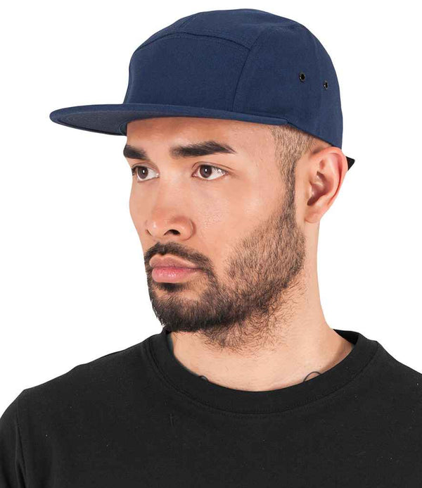 A man with a trimmed beard and mustache wears the Flexfit Classic Jockey Cap by Flexfit in navy blue, featuring a flat peak and adjustable strap, paired with a black crew neck shirt. He faces slightly left against a plain white background.