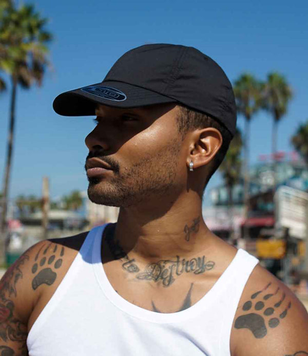 A man wearing the Flexfit 110 Curved Visor Snapback Cap by Flexfit and a white tank top stands outdoors on a sunny day. He has a trimmed beard, tattoos on his neck and shoulders, a hoop earring, with palm trees and blue sky behind him.