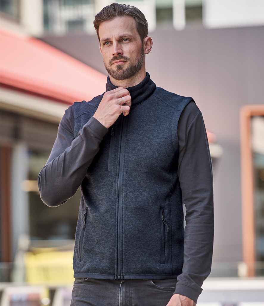 A man with short hair and a beard stands outdoors, adjusting the collar of his Stormtech Avalante Full Zip Knitted Fleece Gilet over a long-sleeve shirt, with an urban building visible in the background.