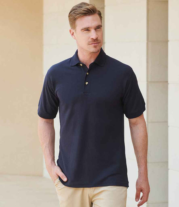 A man with light brown hair stands outdoors in front of a light-colored building, wearing beige pants and a navy Henbury Ultimate Poly/Cotton Piqué Polo Shirt with horn effect buttons by Henbury, one hand in his pocket.